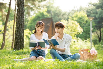 A young male and female couple are enjoying a pleasant picnic while drinking drinks and reading books on the forested park lawn.