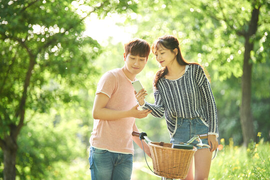 A young male and female couple is riding a bicycle in the park, taking pictures with their smartphones, listening to music, and taking a pleasant walk.