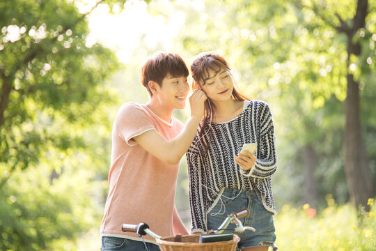 A young male and female couple is riding a bicycle in the park, taking pictures with their smartphones, listening to music, and taking a pleasant walk.