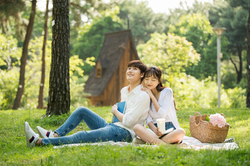 A young male and female couple are enjoying a pleasant picnic while drinking drinks and reading books on the forested park lawn.