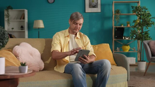 A man using a tablet while sitting on the couch at home close up. A man scrolls through his feed on social networks, looks at photos, videos, laughs. Concept of vacation, relaxation at home.