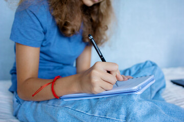 A teenage girl makes notes in her notebook sitting on the bed in the bedroom. Learning process at home.
