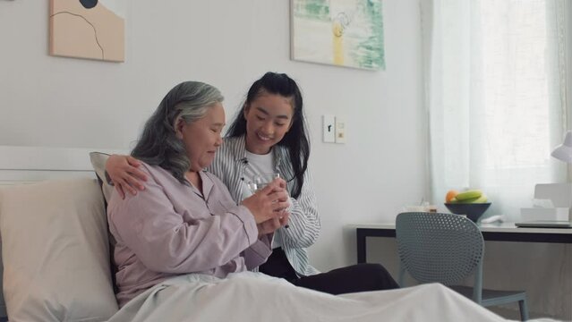 Medium shot of sick mature woman taking glass of water from her young daughter, resting in bed in nursing home