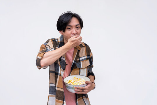 A young asian man voraciously stuffing his mouth with popcorn while watching a movie. Isolated on a white background.