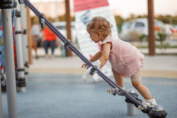 Side view of a 1-3 year old girl climbing a climbing net, rope net or cobwebs on the playground. Toddler with a cast on his arm after playing in the park. White bandage on the arm, plaster on the arm - obrazy, fototapety, plakaty