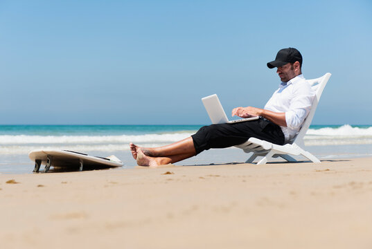 A Businessman Sits On A Beach Chair On The Beach Working On A Laptop Computer With His Surfboard Sitting At His Feet; Tarifa, Cadiz, Andalusia, Spain