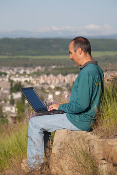 Man Typing On A Laptop Computer Sitting On A Rock Ledge Overlooking A Subdivision With Mountains In The Background; Calgary, Alberta, Canada
