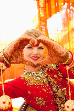 A Bride Wearing An Ornate Gown And Jewelry Making A Heart Shape With Her Hands Covered In Mehndi; Ludhiana, Punjab, India