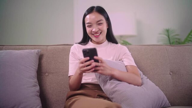 Happy young asian woman relax on comfortable sofa at home texting messaging on smartphone with friend or shopping online from home in her living room at home.