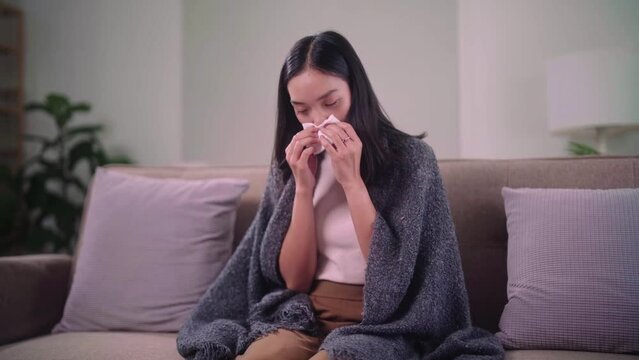 Female unhealthy Sickness, asian young woman has runny and common cold has flu sneezing in a tissue sitting on couch in living room at home, medical and health care concept.
