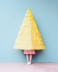 A girl dressed in a pink dress and white shoes carries a large decorated yellow Christmas tree that covers her face, standing in front of a minimal pastel blue wall. Generative AI.