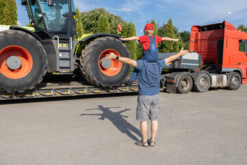 man and boy stand in front of large tractor standing on truck trailer, rear view. son's passion for...
