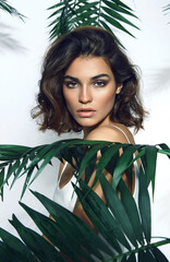 Beautiful young girl in the studio on a white background with dark skin, brown eyes and dark short hair holds a large green tropical leaf in her hands and covers part of her face