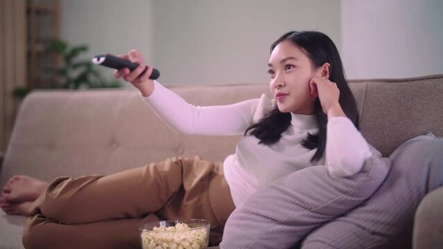 Young asian woman with remote control watching tv and eating popcorn while sitting on the big comfortable sofa in living room at home