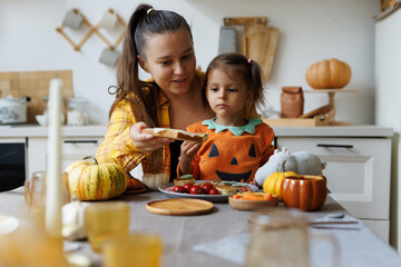 Happy Halloween! Little cute girl and mom eating in the kitchen. A little girl in a pumpkin costume eats sandwiches for breakfast with her mother. Morning at home. - obrazy, fototapety, plakaty