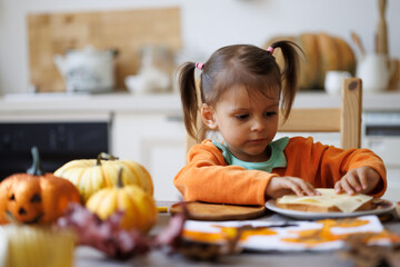 Fototapeta premium Happy Halloween! Little cute girl eating in the kitchen. Toddler girl in a pumpkin costume eating sandwiches on breakfast. Morning at home.