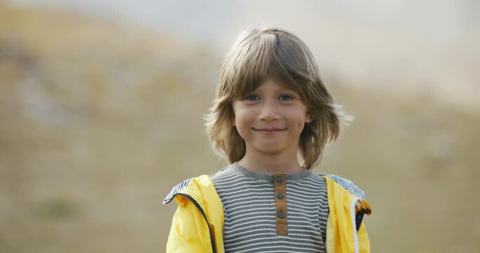 5-6 years old outside. Close-up portrait of child with long hair. Blond-haired and blue-eyed boy looks into frame at first without a smile, then smiles with a charming carefree smile. Cinematic