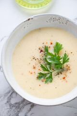 Bowl of cheese cream-soup served with fresh parsley, vertical shot on a white marble background, elevated view, close-up