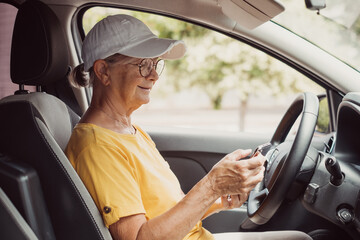 Fototapeta na wymiar Senior woman stops driving the car to use smartphone, elderly female satisfied with unforgettable journey by car, sits on driver`s seat. People, driving, transport concept
