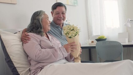 Medium shot of Asian cheerful mature husband giving bouquet of flowers to his sick wife resting in bed at home - Powered by Adobe