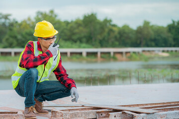 An engineer wearing a yellow hat, red shirt and green outerwear carries a radio at the construction site.