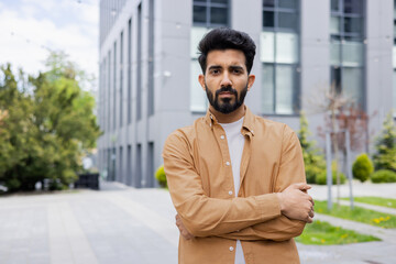 Serious thinking businessman programmer looking at camera with crossed arms, indian man focused outside office building in shirt.