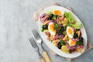 Tuna salad with pasta, eggs, potatoes, olives, red onions and sauce in white plate on old light gray concrete table background. Nicoise salad. French cuisine. Top view. Flat lay.