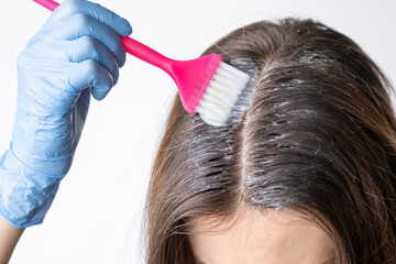 Fototapeta premium Close-up of a woman's heClose-up of a woman's head in the process of hair coloring on a white background. Closeup woman hands dyeing hair using a brush. Colouring of white hair at home.