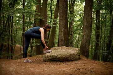 Woman runner stands near large stone and tying her shoelaces, ready to run through forest
