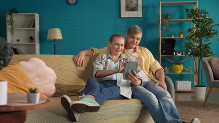 A man and a woman look at photos and videos on a tablet. Portrait of a married couple sitting in an embrace on the sofa in the living room. Family idyll.
