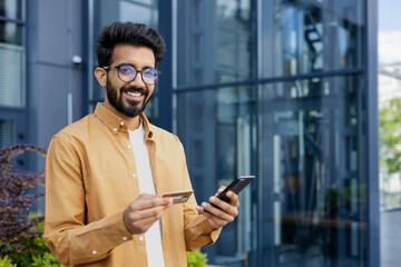 Portrait of young man outside office building, hispanic smiling and looking at camera, holding...