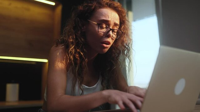 Nervous stressed curly woman freelancer or student with glasses typing quickly on laptop computer and looking at screen asking to hurry up scream and warning about deadline at home