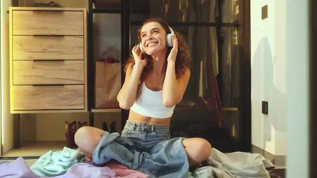 Funny happy curly woman with headphones sitting on clothes and dancing while cleaning apartment at home Cute expressive female enjoying housekeeping indoors alone