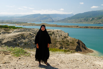A Muslim woman in a black niqab stands on top of a mountain against the backdrop of a mountain lake.