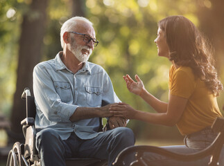 Senior man in wheelchair and daughter talking and smiling in park - 647781696