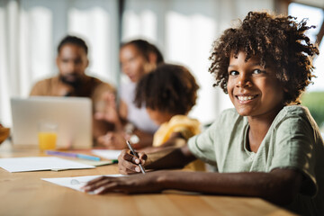 Happy African American boy coloring on notepad and looking at camera while being with his family