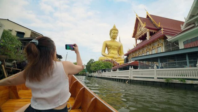 Young Asian woman tourist sitting on boat using smartphone taking photo of the giant buddha statue at the Wat Paknam Phasi Charoen temple in Bangkok, Thailand. Travel and transport lifestyle