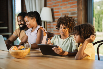 Young black family using digital tablet and laptop at dining table - obrazy, fototapety, plakaty