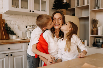 Teenage children kiss a smiling mother sitting at a table in a decorated kitchen interior on a Christmas holiday at home. Family lifestyle. Selective focus