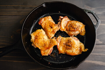 Seared Chicken Thighs in a Cast-Iron Skillet: Four pan-fried chicken thighs with skin in a skillet placed on a wooden table