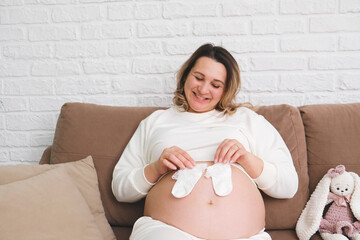 Beautiful pregnant woman is holding baby socks on her belly on sofa in light living room.