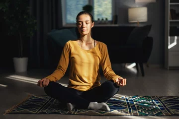 Poster Young woman sitting in lotus pose at home, sun rays on woman's face, concept of inner peace and relax © MYDAYcontent