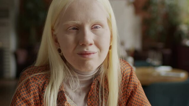 Selective focus close-up portrait of young albino woman with long hair sitting in modern restaurant looking at camera