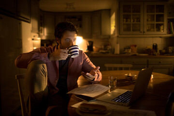 Young Caucasian man drinking coffee while going over bills and payments at night at home