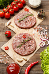 Raw burger patties on parchment and hamburger ingredients on a wooden background.