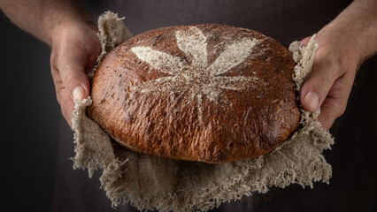Man baker holding fresh spelt wheat hemp flour loaf of bread on rustic linen towel on dark background. Loaf bread  with crushed hemp seeds decorated cannabis leaf from flour.