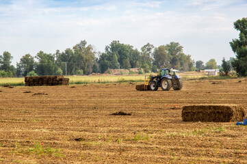 Tractor agrícola amontonando pacas de heno en un campo segado, bajo un cielo azul con nubes.
