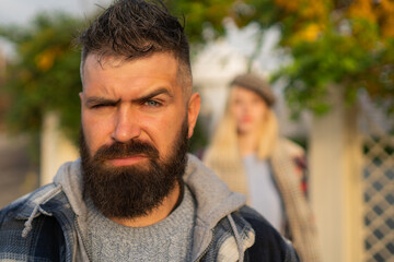 Fototapeta premium Bearded brutal masculine man raises an eyebrow looking at camera with indignation. Beautiful blonde girl standing behind male at blured background. Couple in love had an argue.
