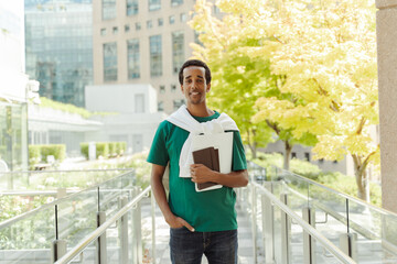 Young smiling African American student holding documents, notebook wearing stylish green t shirt, looking at camera standing outdoors on urban city background. Successful business concept