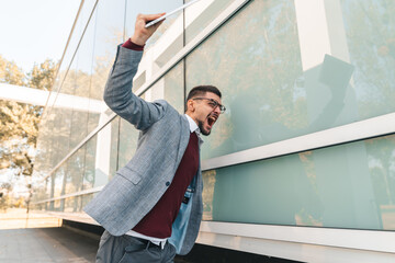 Young angry businessman with glasses yelling outside office building. Male manager under stress feeling furious and frustrated after work.